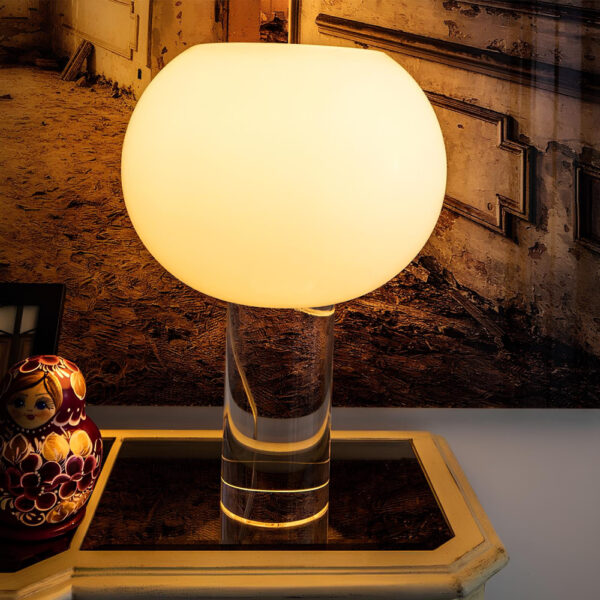 Lampe champignon avec pied cylindrique transparent et boule lumineuse diffusant une lumière blanc chaud. Posée sur un support vintage marron. Un tableau ancien sur le mur derrière.